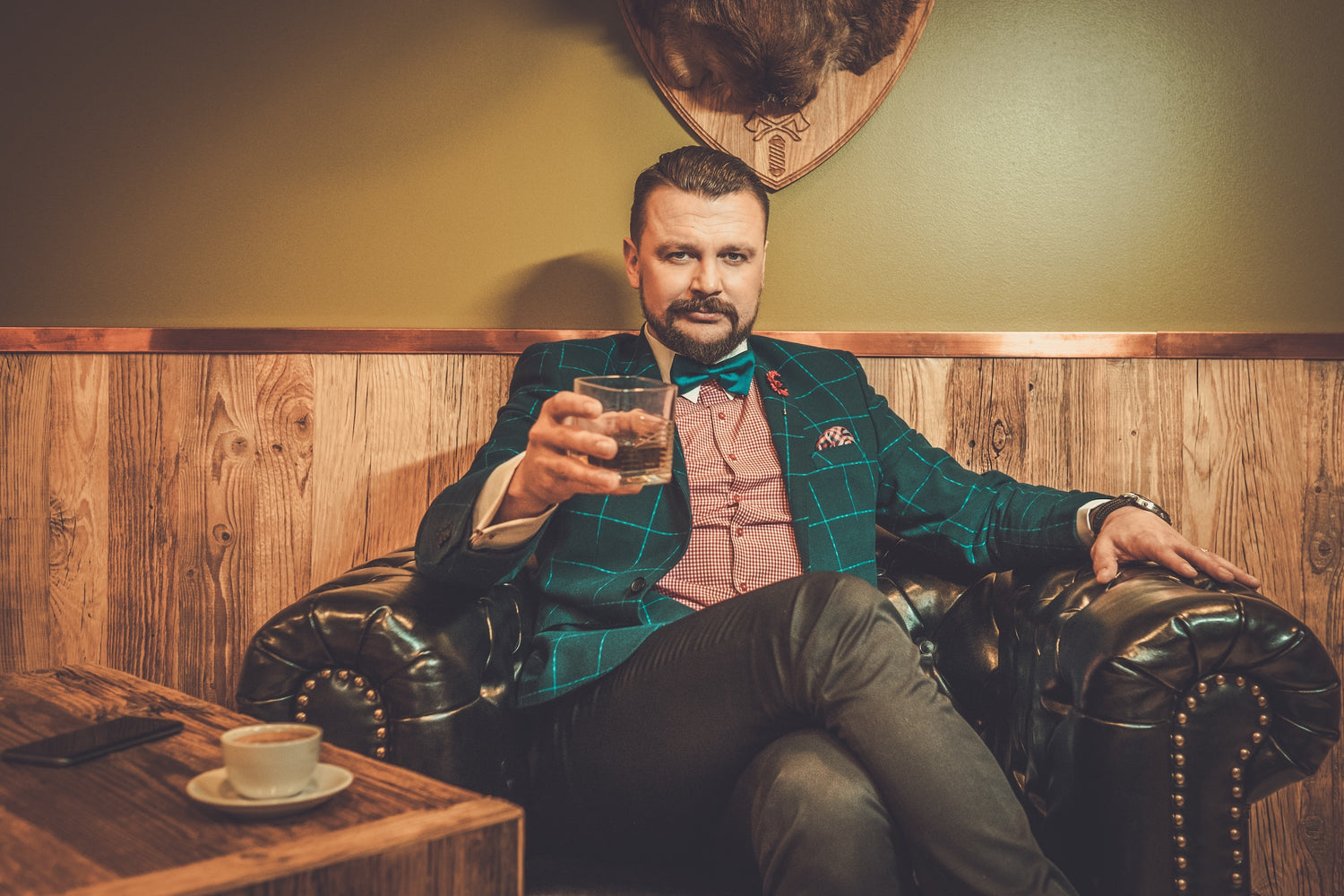 Well-dressed bearded man, adorned in a green windowpane sportcoat, elegantly seated in a leather chair. Behind him, the wall is adorned with a deer euro mount, adding to the room's sophisticated ambiance. He sports an inviting smirk and confidently extends a glass of scotch towards the viewer, capturing a moment of camaraderie as if proposing a toast or cheers.