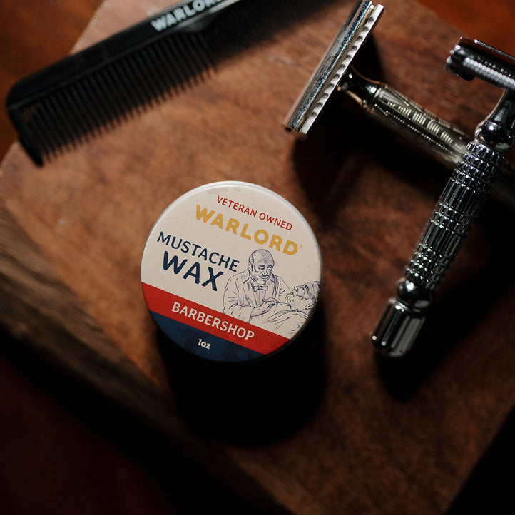 Barbershop Mustache Wax by Warlord next to straight razor and comb on rustic wooden table.