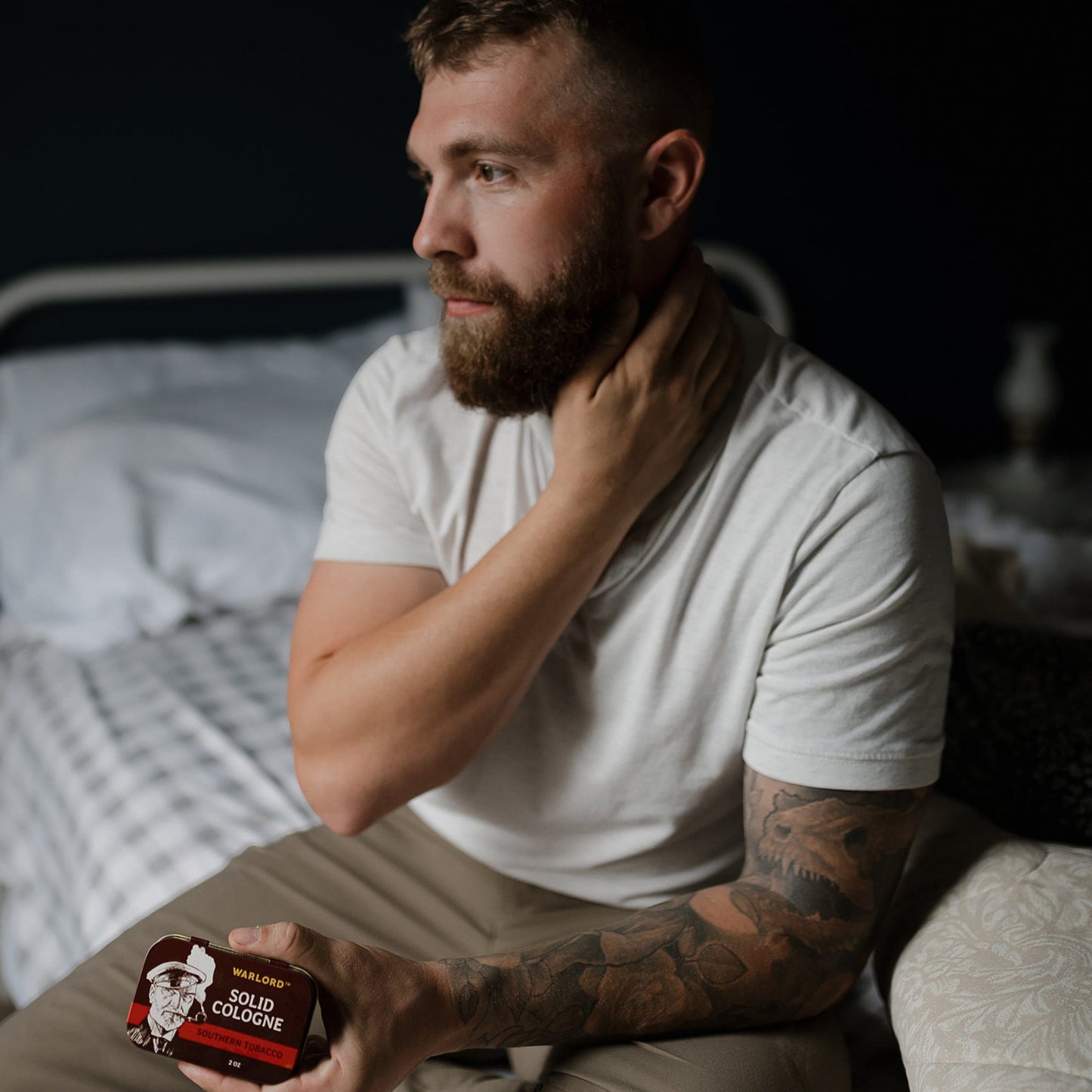 Bearded man holding Warlord Southern Tobacco solid cologne tin on bed with tattooed arm - Photograph