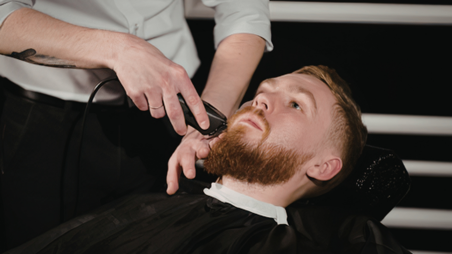 Barber trimming a man's red goatee into shape in a traditional barbershop chair.