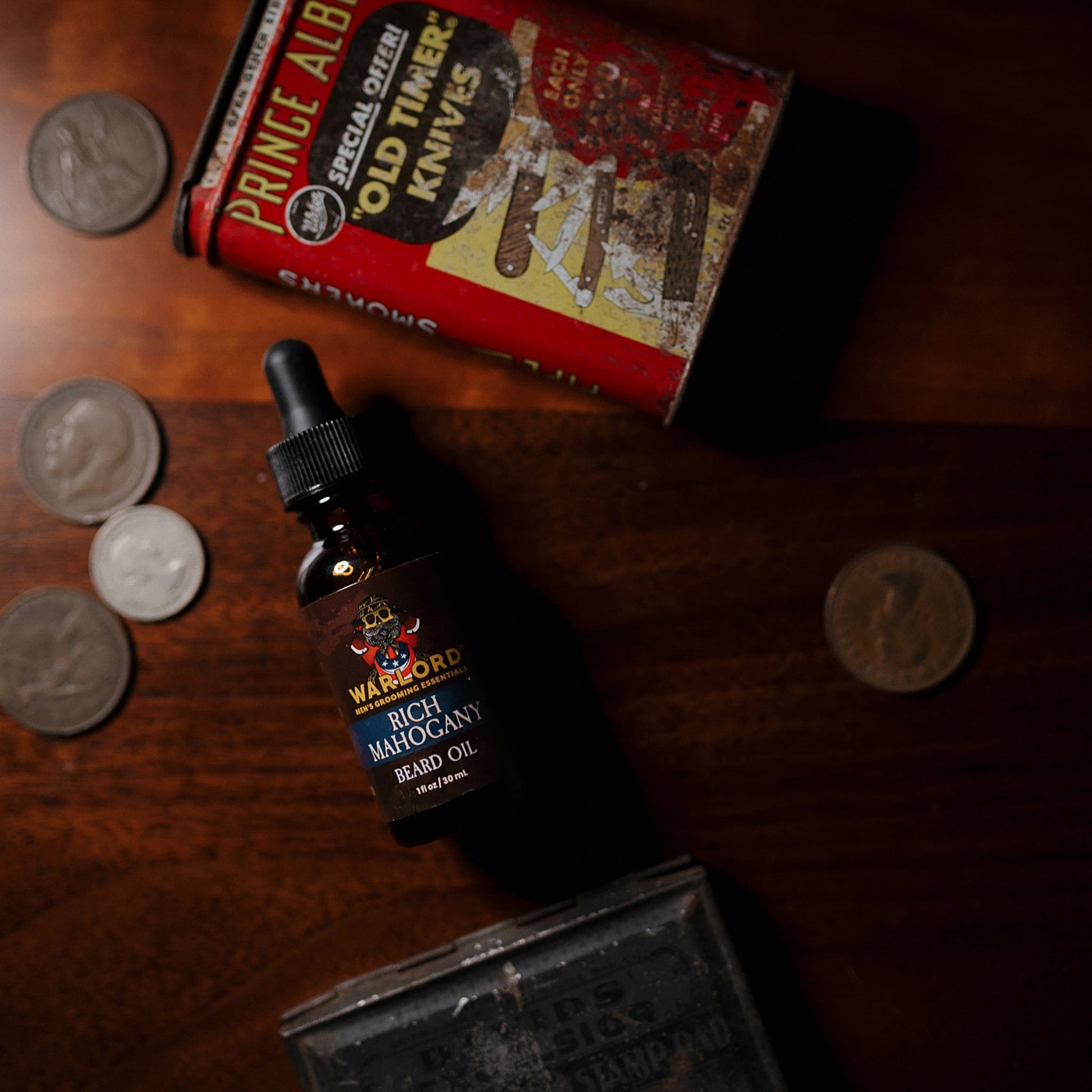 Warlord Rich Mahogany Beard Oil bottle placed on a wooden surface surrounded by vintage items including coins, a Prince Albert tobacco tin, and an old knife box—evoking a nostalgic, rugged vibe.
